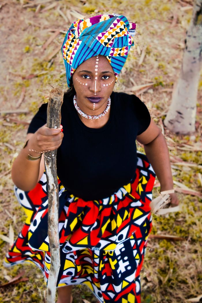 Vibrant image of an African woman in traditional attire with cultural face paint and stick, standing outdoors.